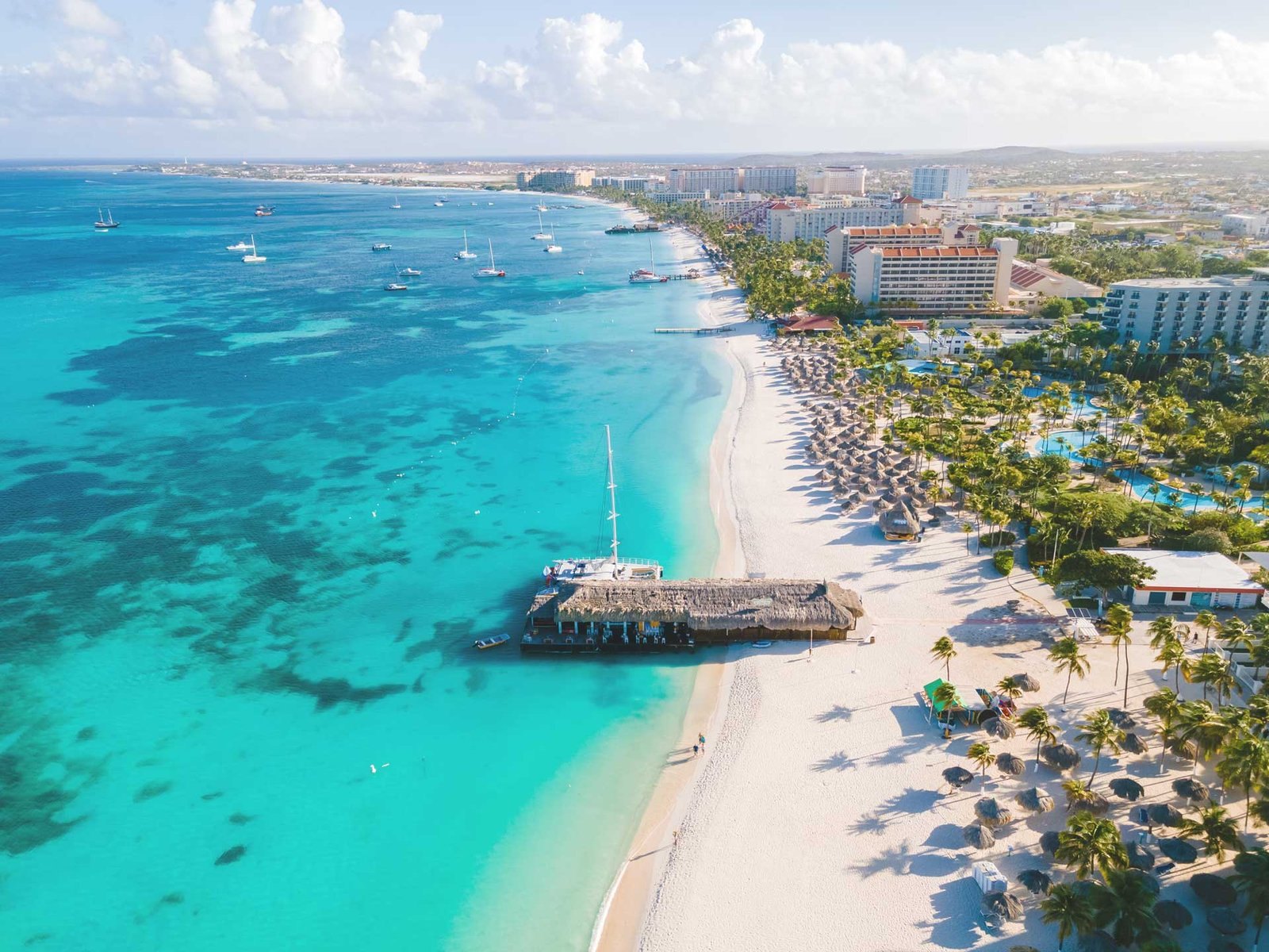 Aerial photograph of Palm Beach, Aruba, with palapas lining the sand below the high-rise hotel strip; YOO Aruba's site in Ponton lies about 2.7 miles away.